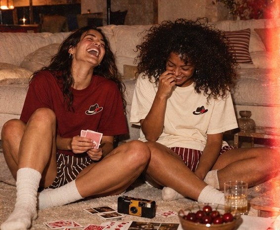 Two friends laughing over a hand of cards on the floor, cherries in a bowl nearby
